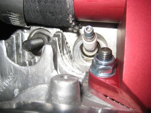 At the rear location, the screw is barely visible behind and to the left of the spark plug.